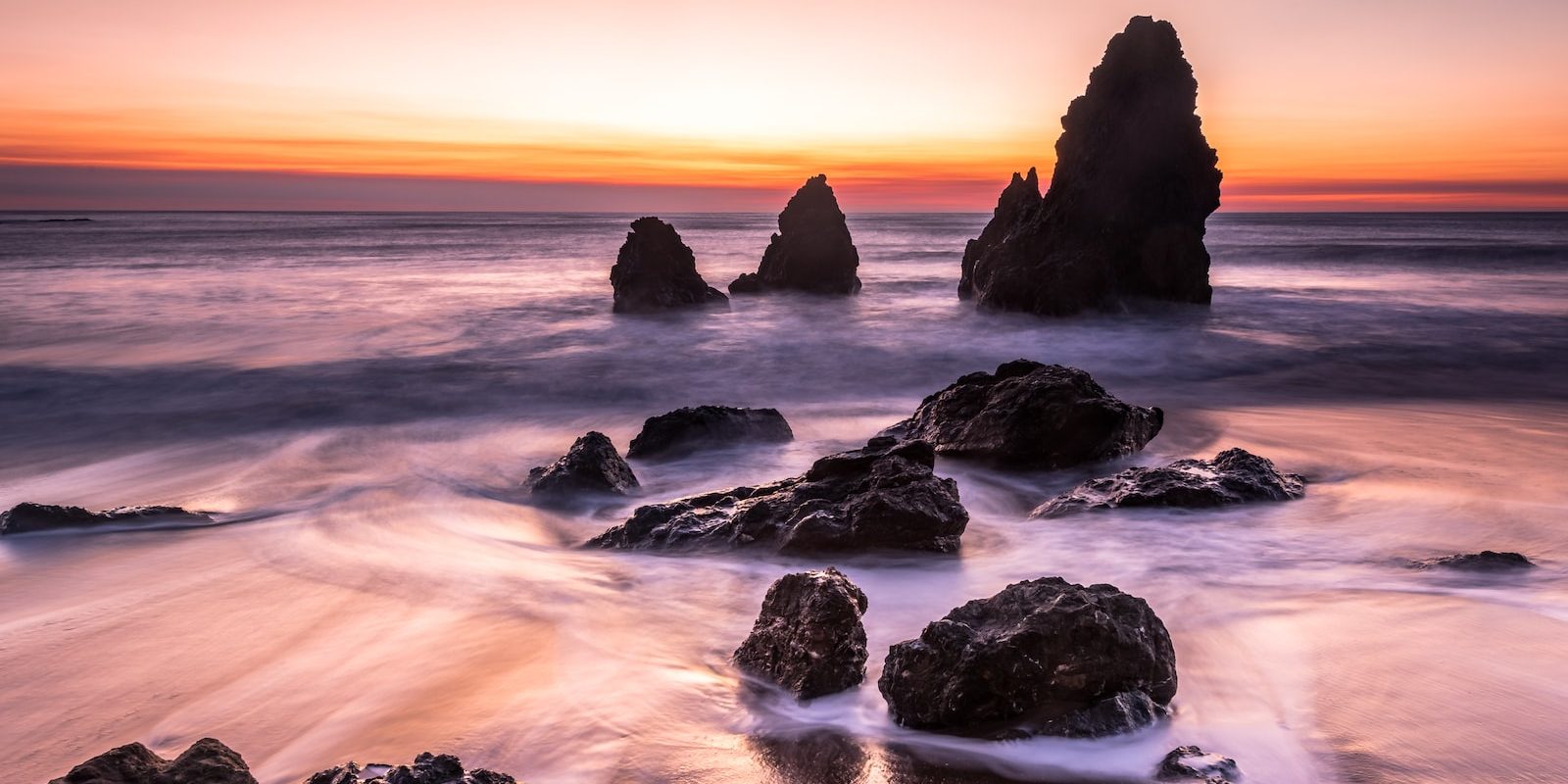 photography of beach near black rock formation under orange sky